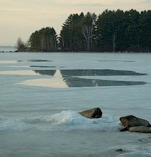 Snow and ice covered waterbody with some open patches of water. Woodlands in the background.