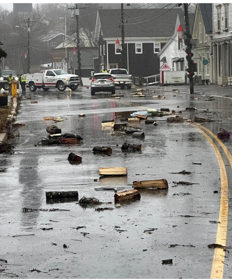 Debris on a flooded roadway with cars and gray buildings. 